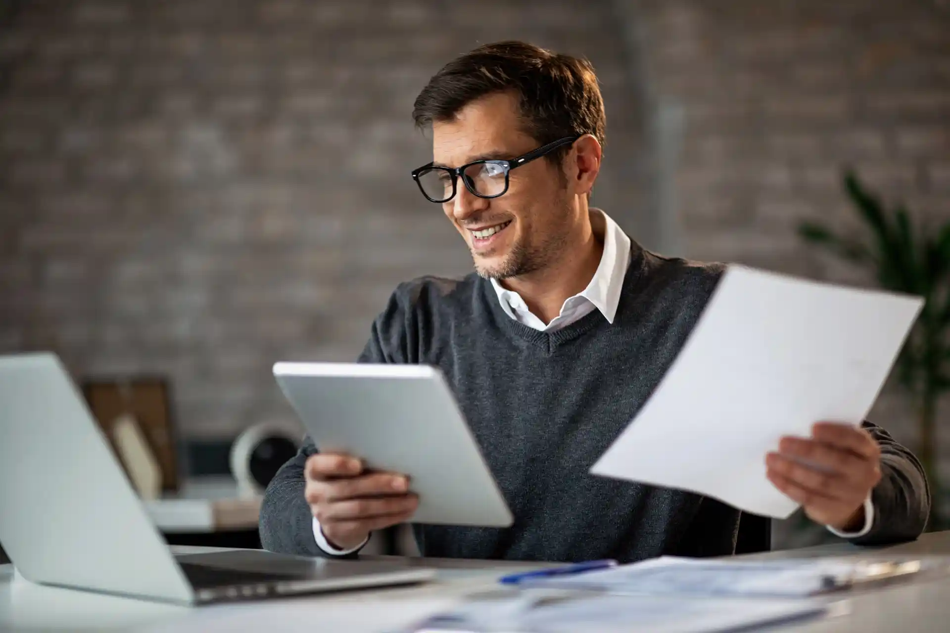 Mann mit Brille arbeitet am Schreibtisch mit Laptop, Tablet und Dokumenten.