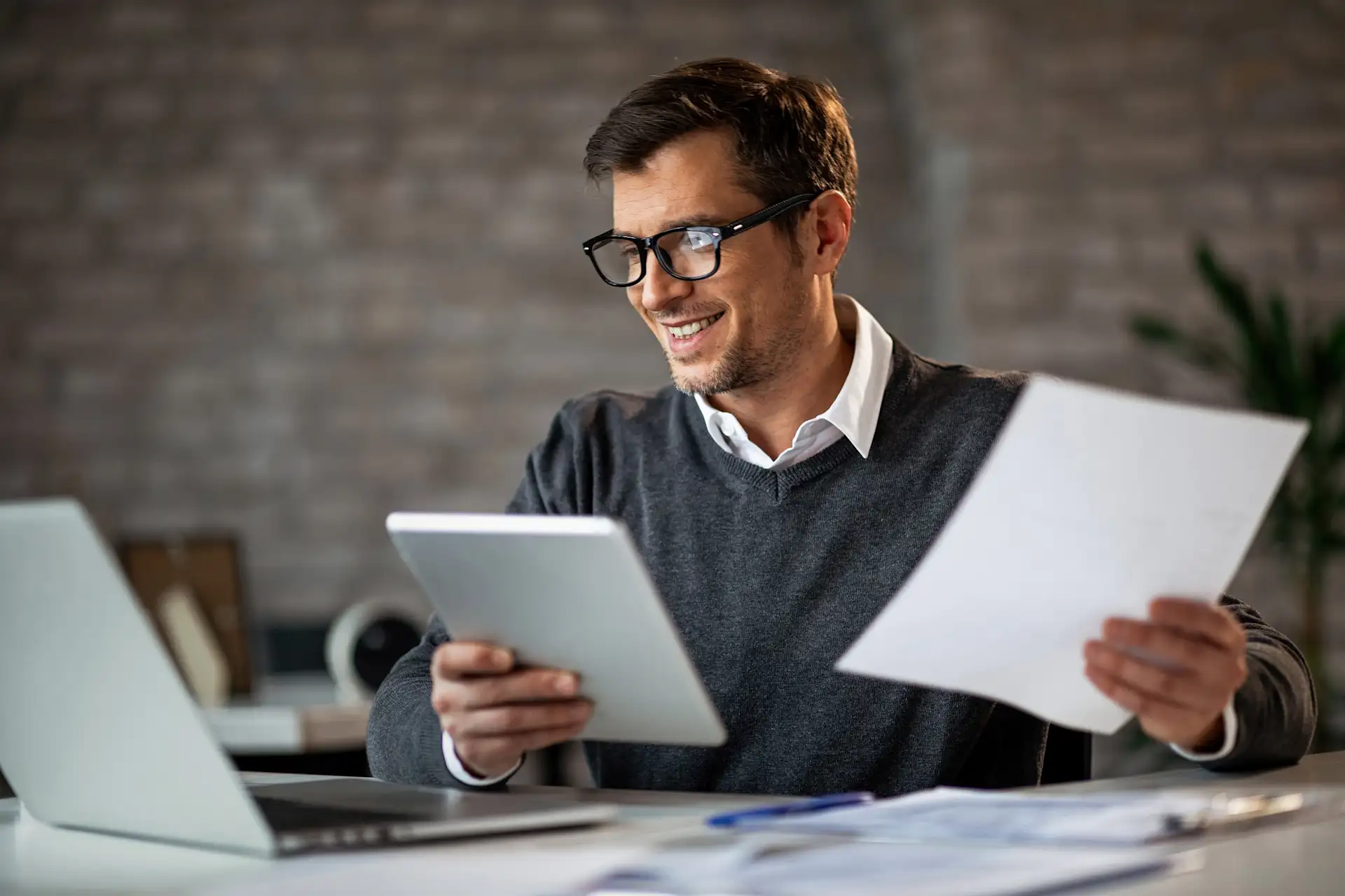 Mann mit Brille arbeitet am Schreibtisch mit Laptop, Tablet und Dokumenten.
