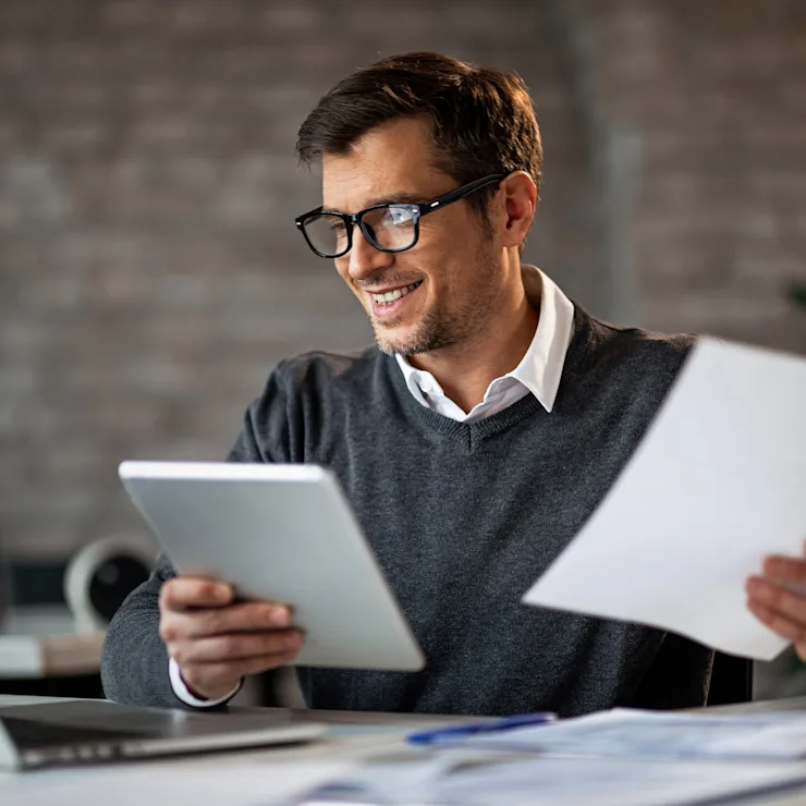 Mann mit Brille arbeitet am Schreibtisch mit Laptop, Tablet und Dokumenten.