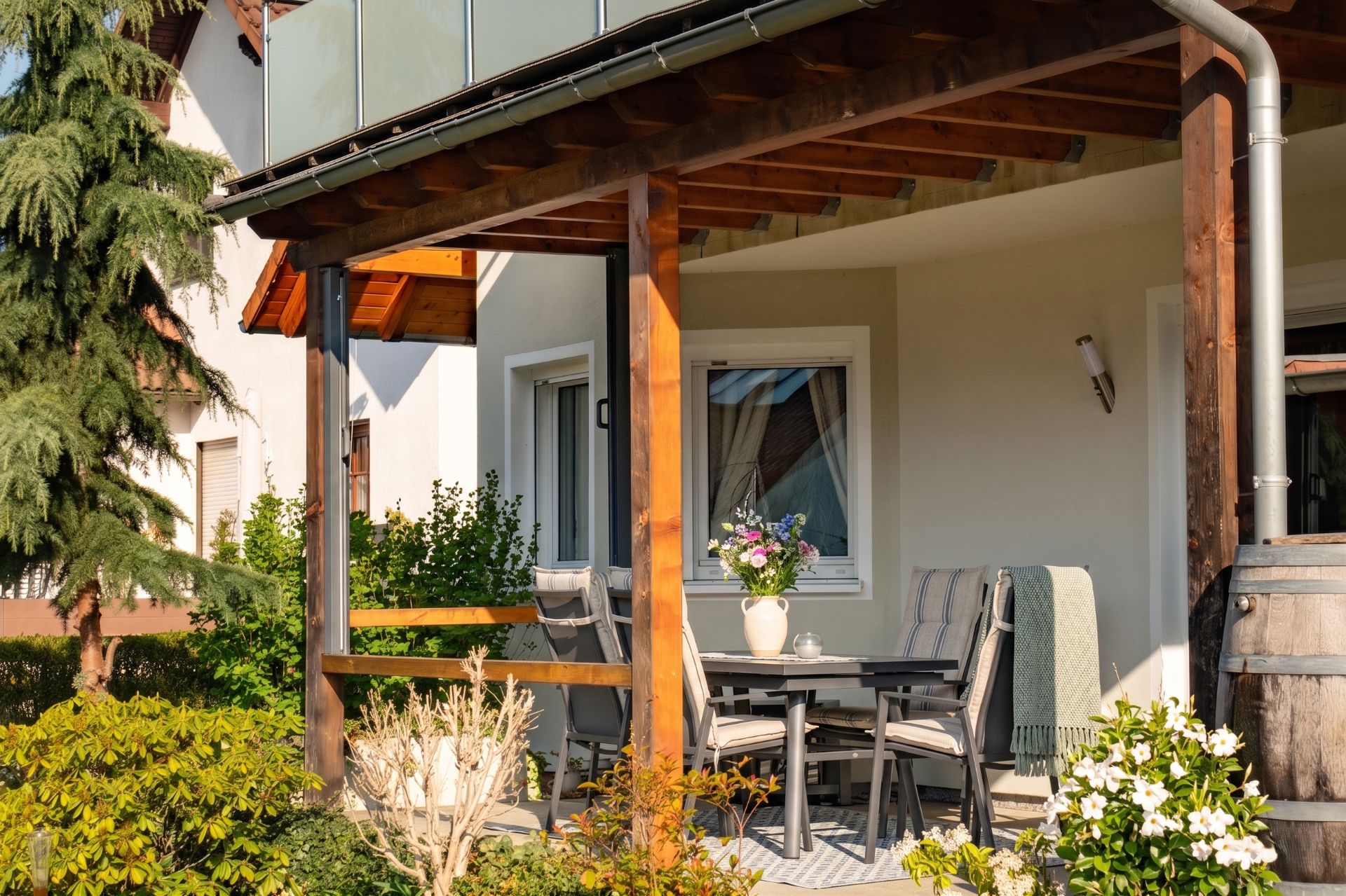 Überdachte Terrasse mit Tisch, vier Stühlen und einer Blumenvase, umgeben von Pflanzen.