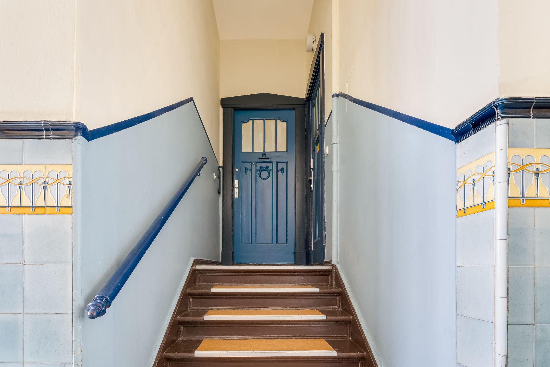 Treppenaufgang mit blauer Tür und hellblau gefliesten Wänden mit gelbem Muster.