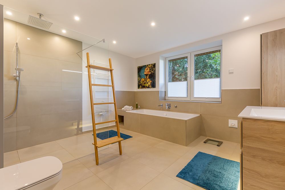 Modernes Badezimmer mit begehbarer Dusche, Badewanne, Holzleiter, Fenster und blauen Badvorlegern.