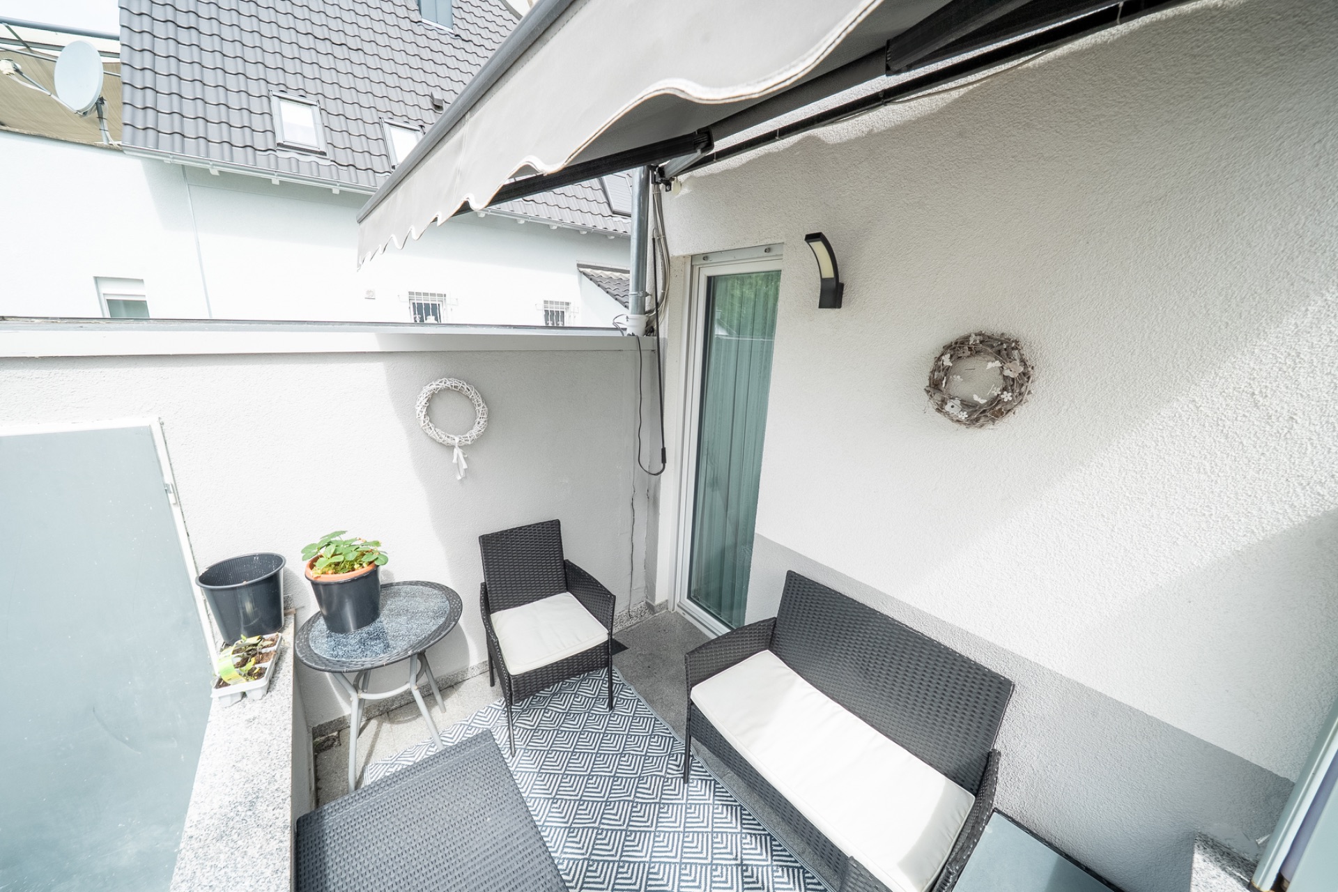 Kleine Terrasse mit Sitzmöbeln, Tisch, Pflanzen und Wanddekoration unter einer Markise.