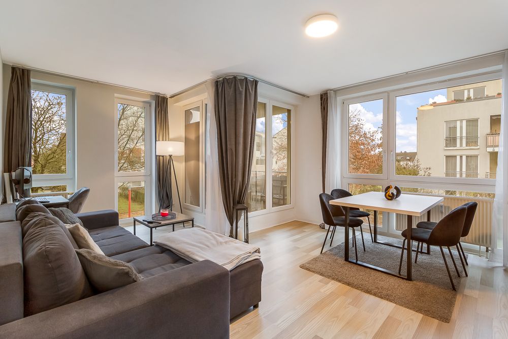 Helles Wohnzimmer mit grauem Sofa, Esstisch für vier Personen und großen Fenstern.