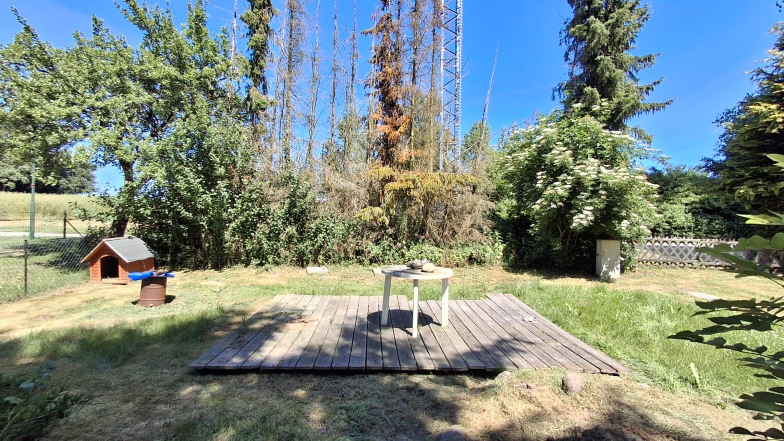 Garten mit Holzterrasse, rundem Tisch und Hundehütte, umgeben von Bäumen und Sträuchern.