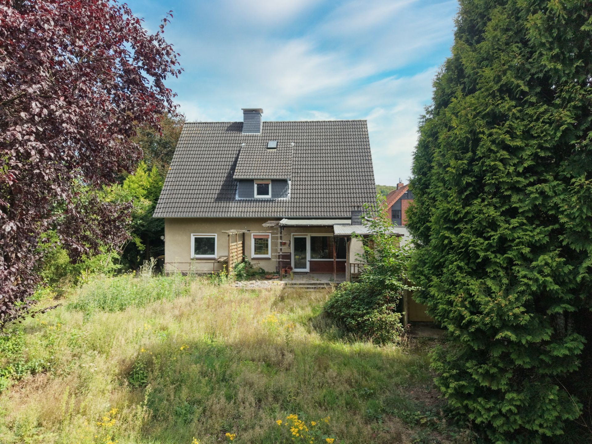 Zweistöckiges Haus mit schrägem Dach, umgeben von Bäumen und wildem Garten.