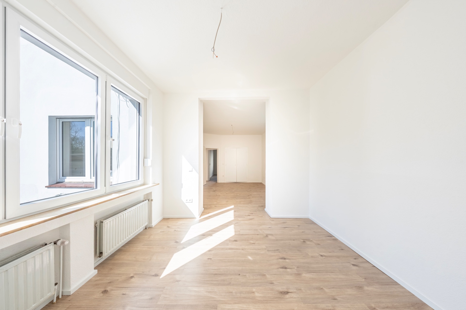 Heller, leerer Raum mit großen Fenstern, hellem Holzboden und weißen Wänden.