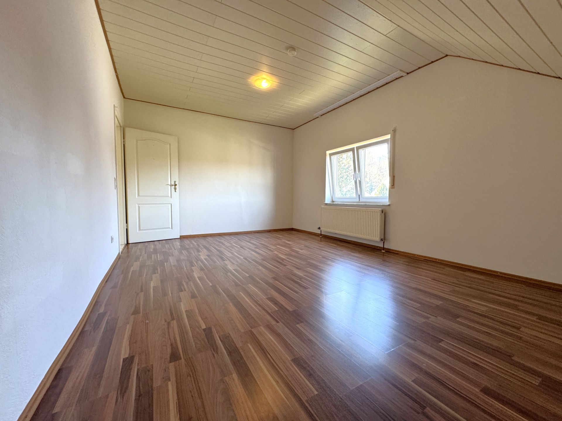 Leeres Zimmer mit Holzfußboden, weißer Tür und Fenster mit Heizkörper darunter.