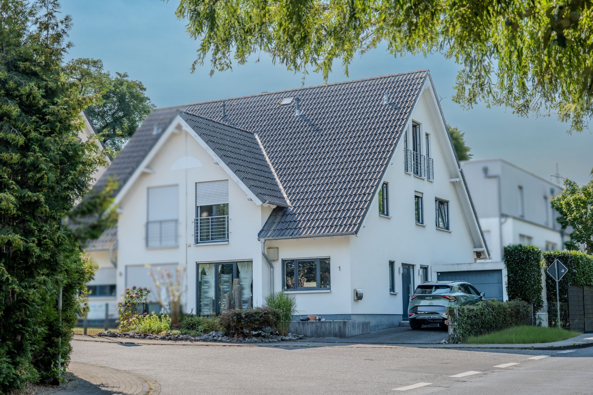 Modernes weißes Wohnhaus mit grauem Dach, großem Fenster und zwei Autos in der Einfahrt.