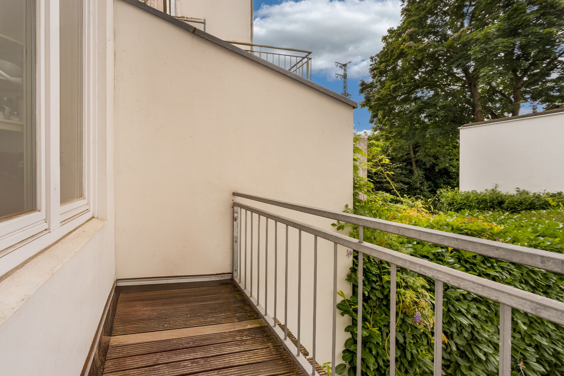 Kleiner Balkon mit Holzboden und Metallgeländer, Blick auf grüne Büsche und Bäume.