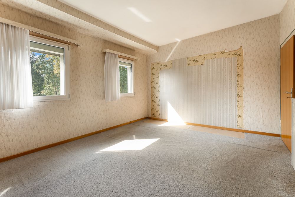 Heller, leerer Raum mit zwei Fenstern, Teppichboden und gemusterter Tapete.