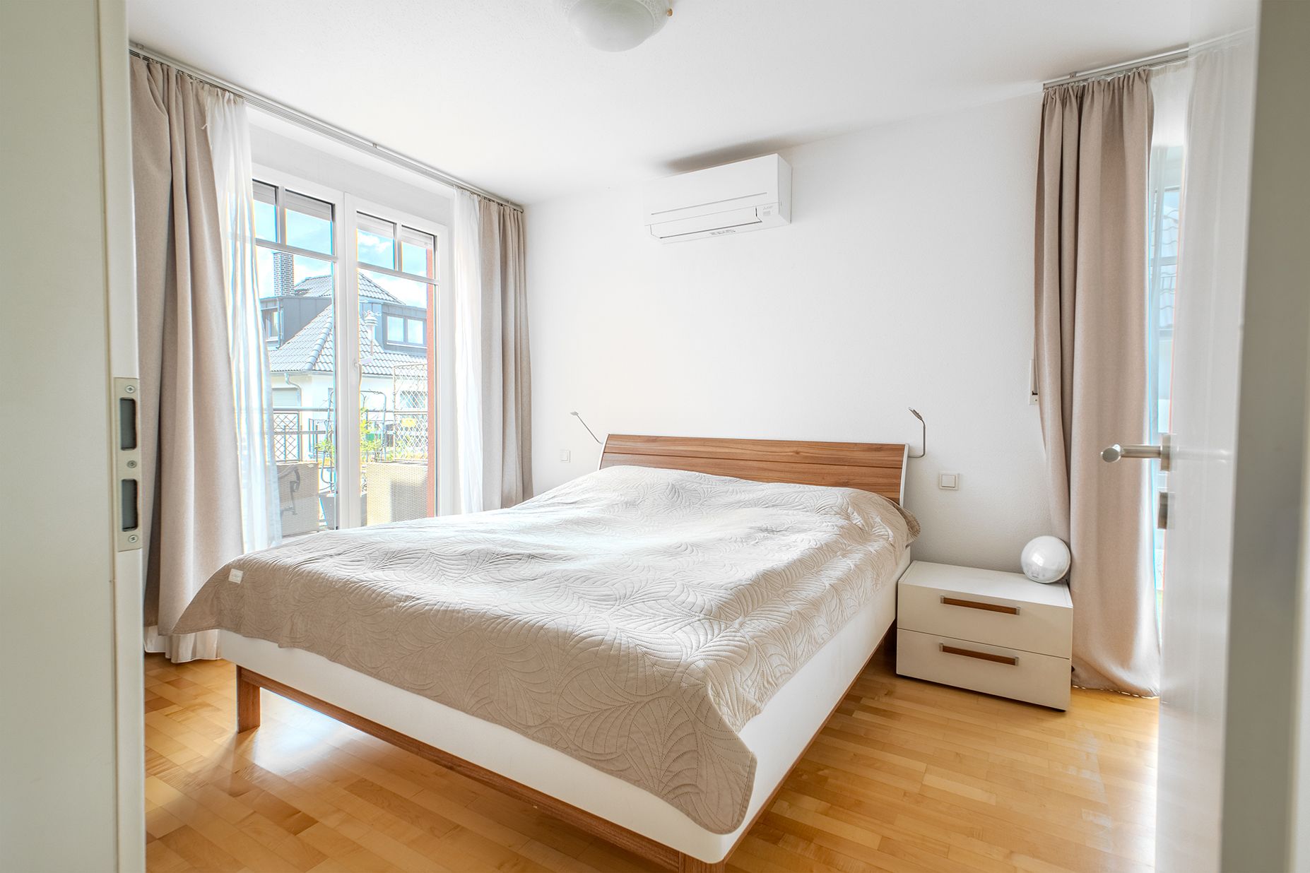 Modernes Schlafzimmer mit Doppelbett, Nachttisch, Klimaanlage und großen Fenstern mit Vorhängen.