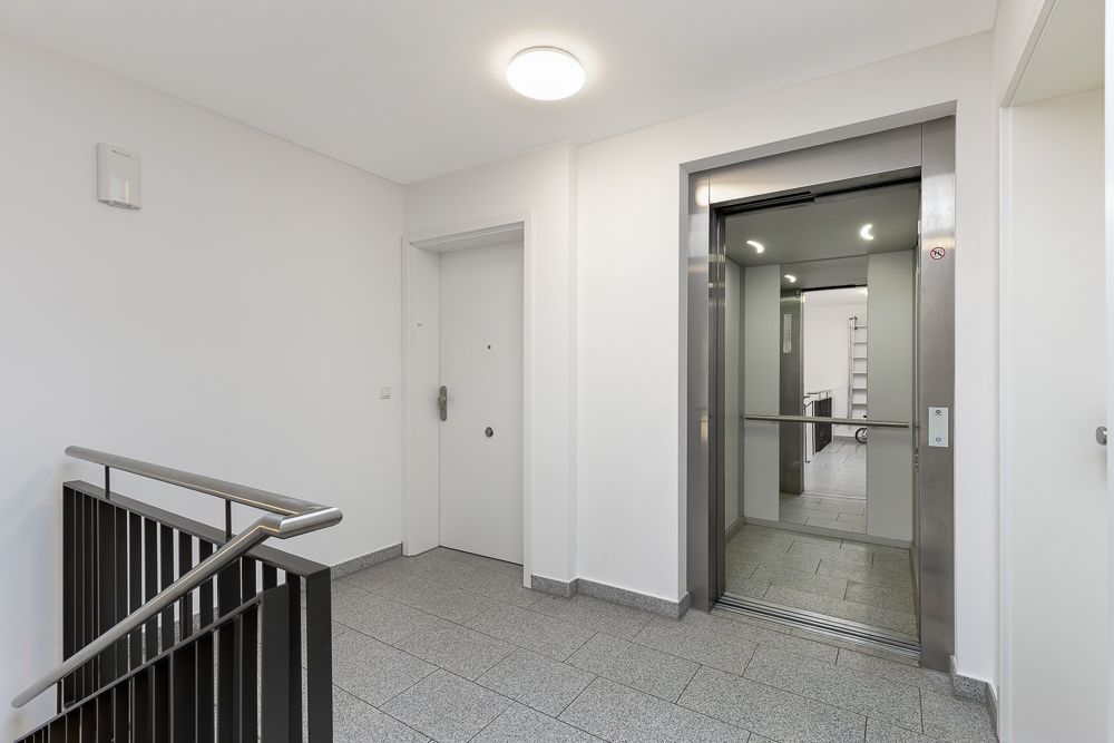 Heller Flur mit Aufzug, Wohnungstür und Treppengeländer.