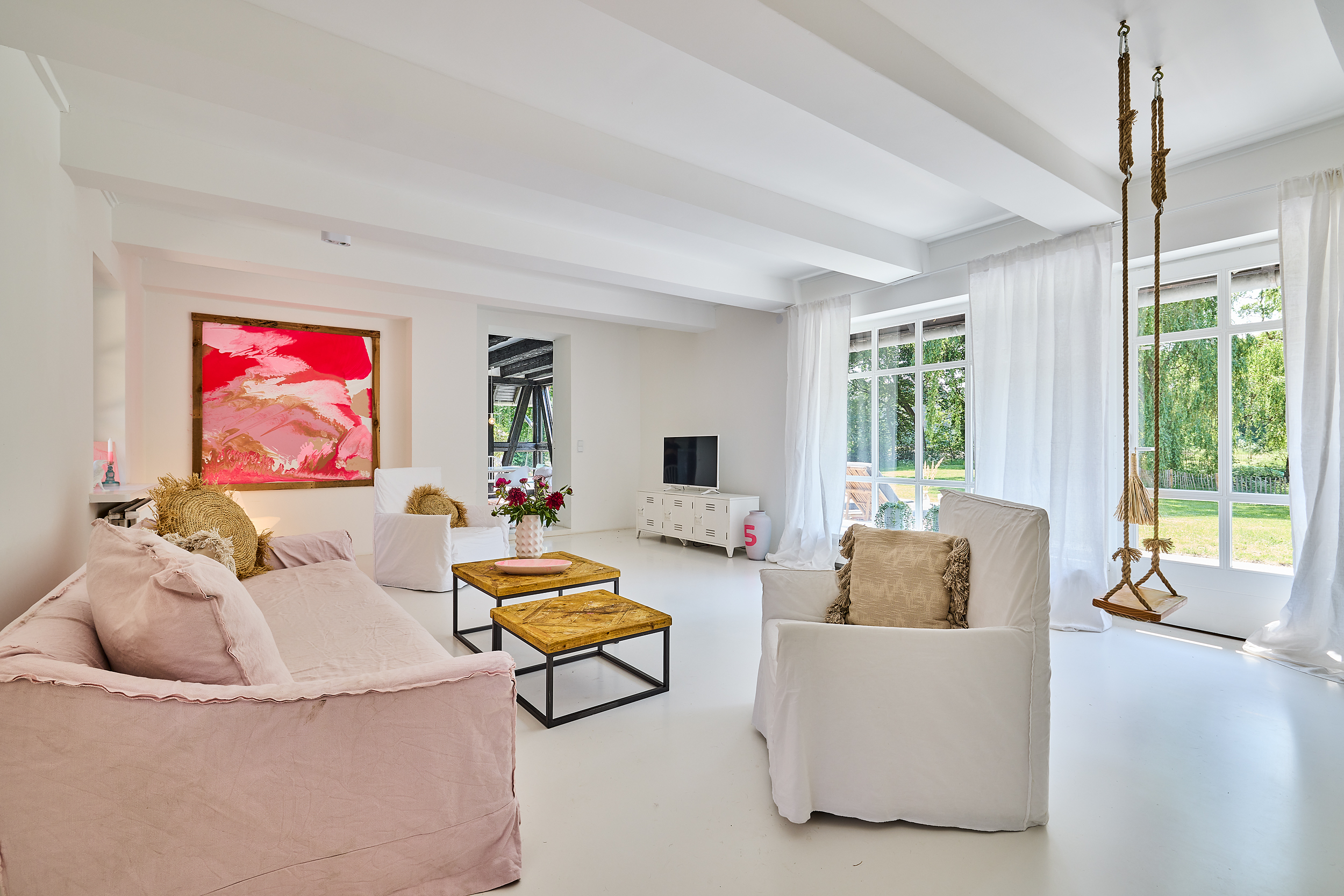 Helles Wohnzimmer mit Sofas, Schaukel, buntem Bild und großen Fenstern zum Garten.