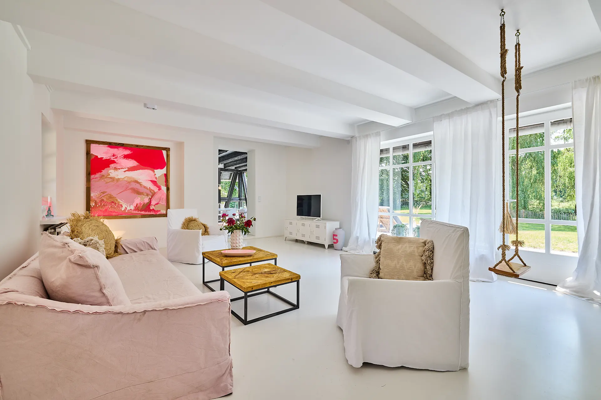 Helles Wohnzimmer mit Sofas, Schaukel, buntem Bild und großen Fenstern zum Garten.