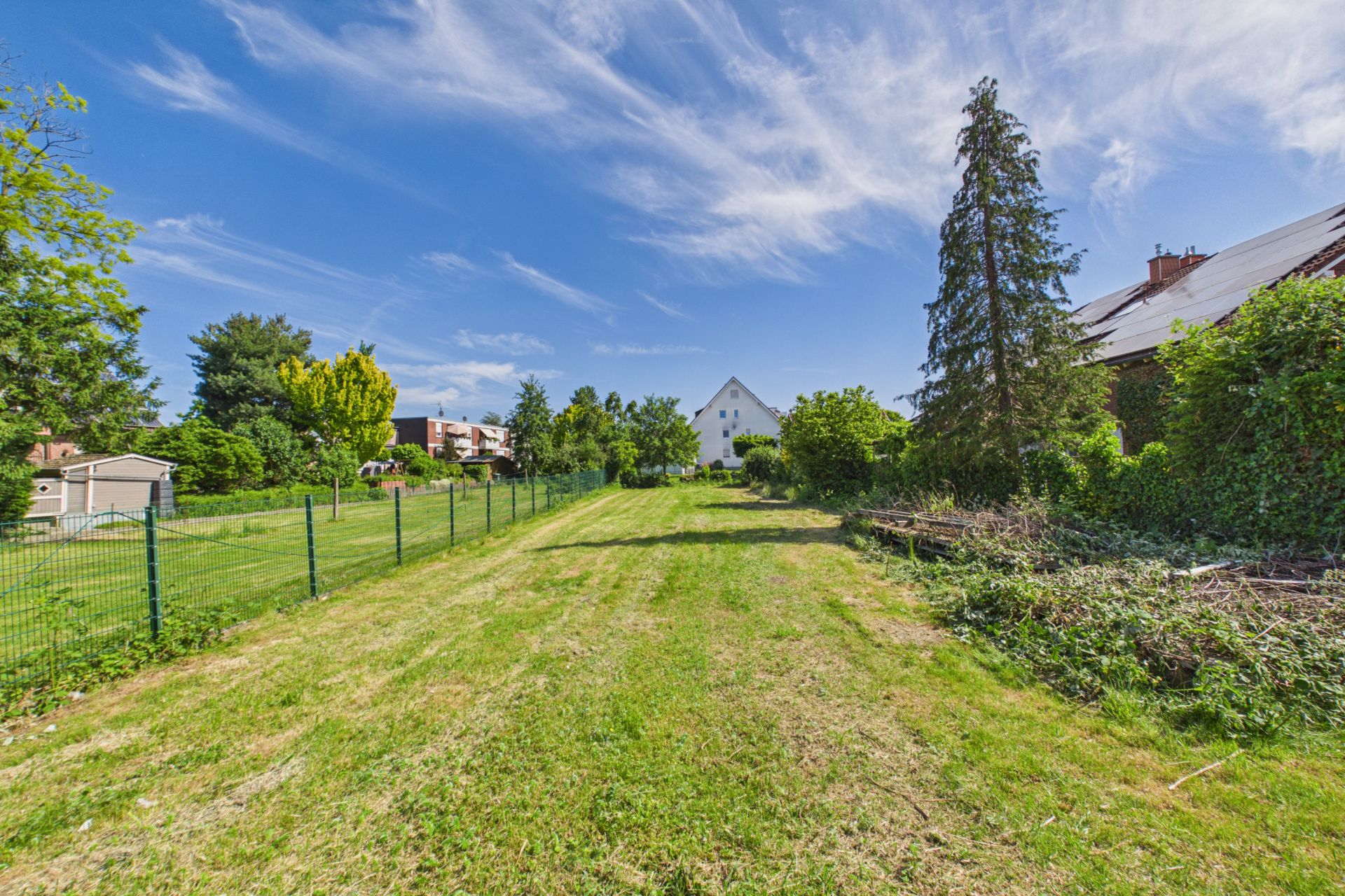 Großer, gepflegter Garten mit Rasen, Bäumen und angrenzenden Häusern bei blauem Himmel.