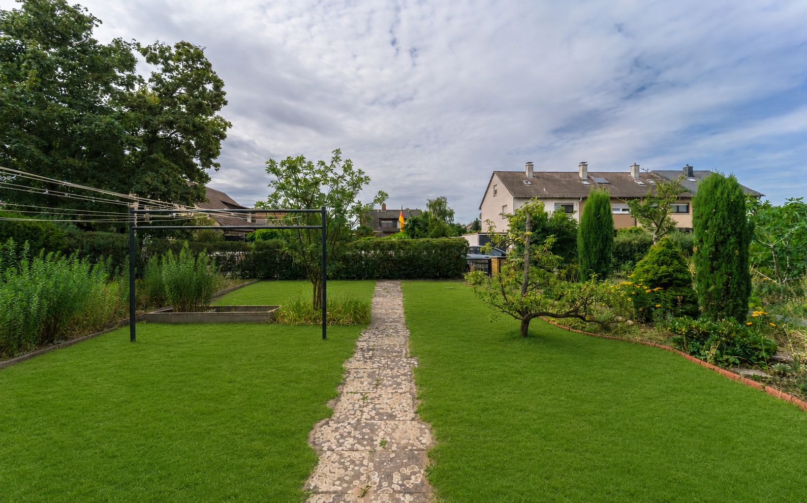 Gepflegter Garten mit Rasen, Steinweg, Bäumen und Sträuchern, umgeben von Wohnhäusern.
