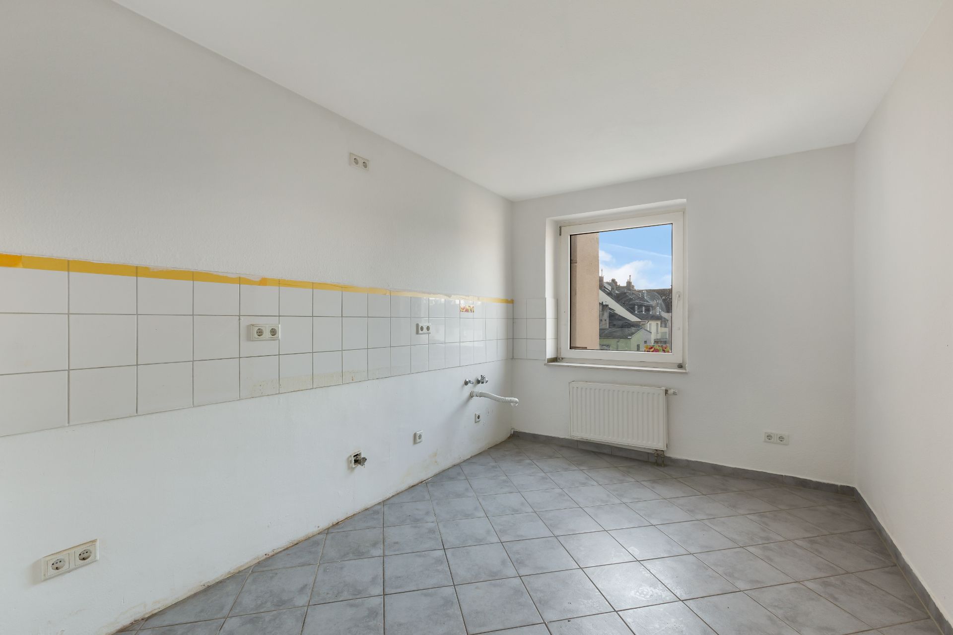 Leerer Raum mit grauen Fliesen, weißen Wänden und einem Fenster, Anschlussstellen für eine Küche sichtbar.
