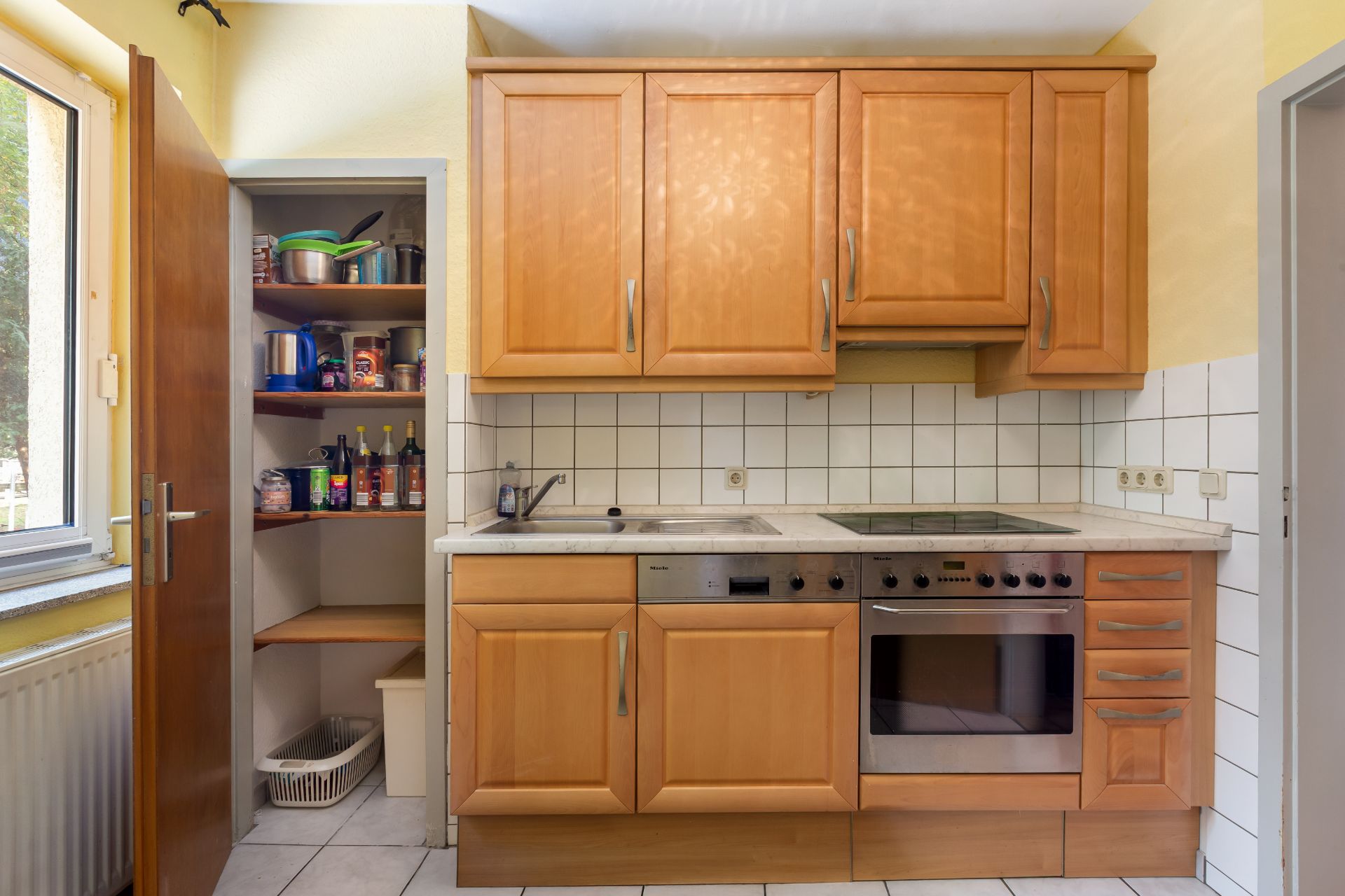 Kleine Küche mit Holzschränken, Herd, Backofen und offener Vorratskammer mit Regalen.