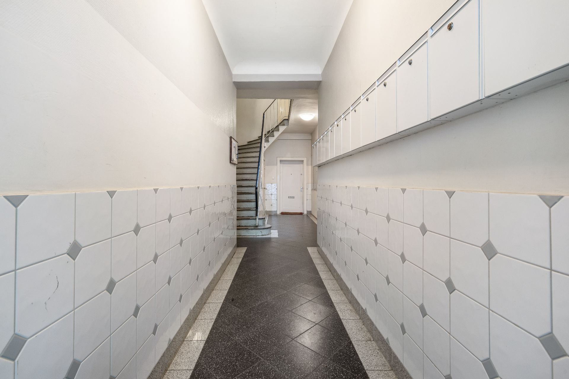 Flur mit Fliesenwänden, Briefkästen an der rechten Wand und Treppe im Hintergrund.