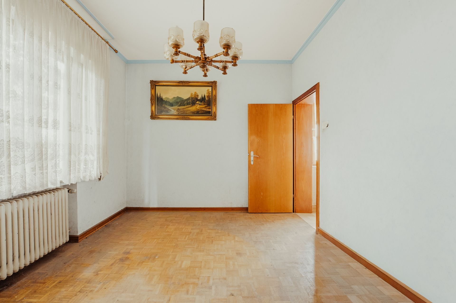 Leerer Raum mit Parkettboden, Kronleuchter, Heizung, Fenster mit Vorhängen und offenem Holztür.