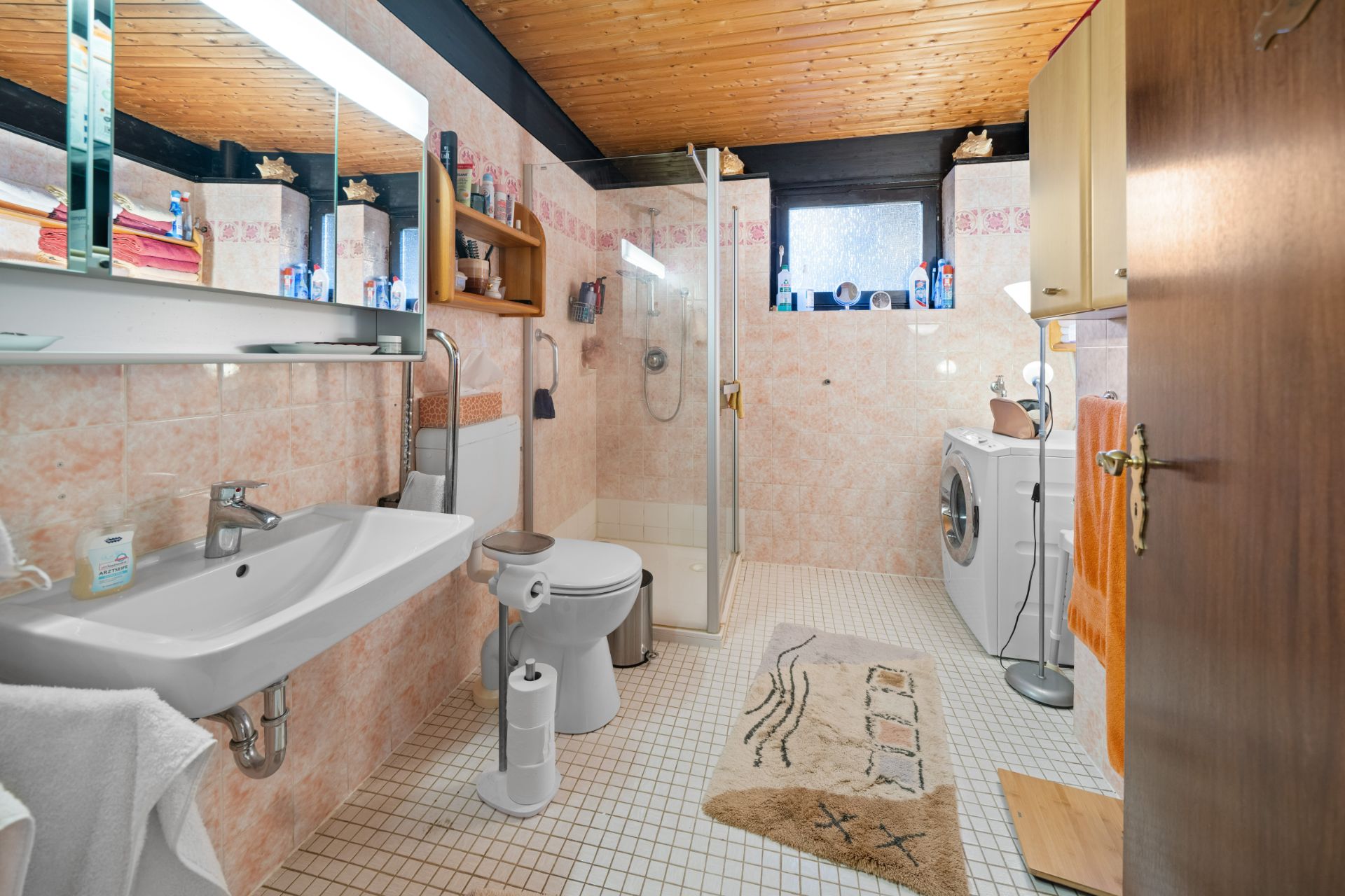 Badezimmer mit Waschbecken, Toilette, Dusche, Waschmaschine und Regal, helle Fliesen und Holzdecke.