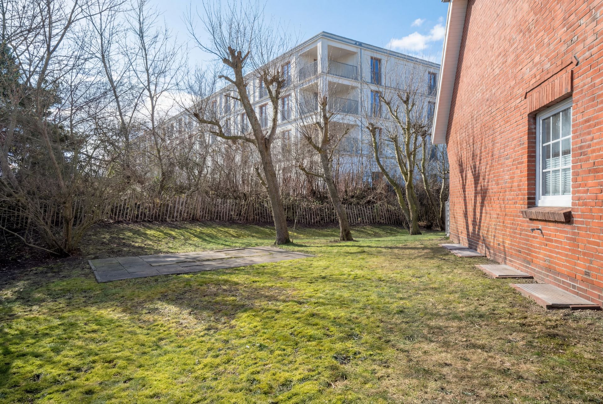 Garten mit Rasen, Bäumen, einem Zaun und roter Hauswand rechts im Bild.