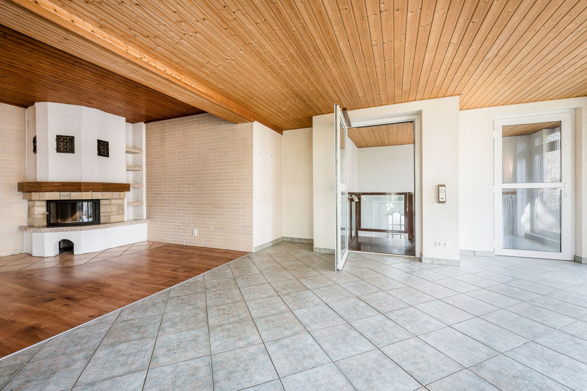 Helles Wohnzimmer mit Kamin, Fliesen- und Parkettboden, offener Tür und Holzdecke.