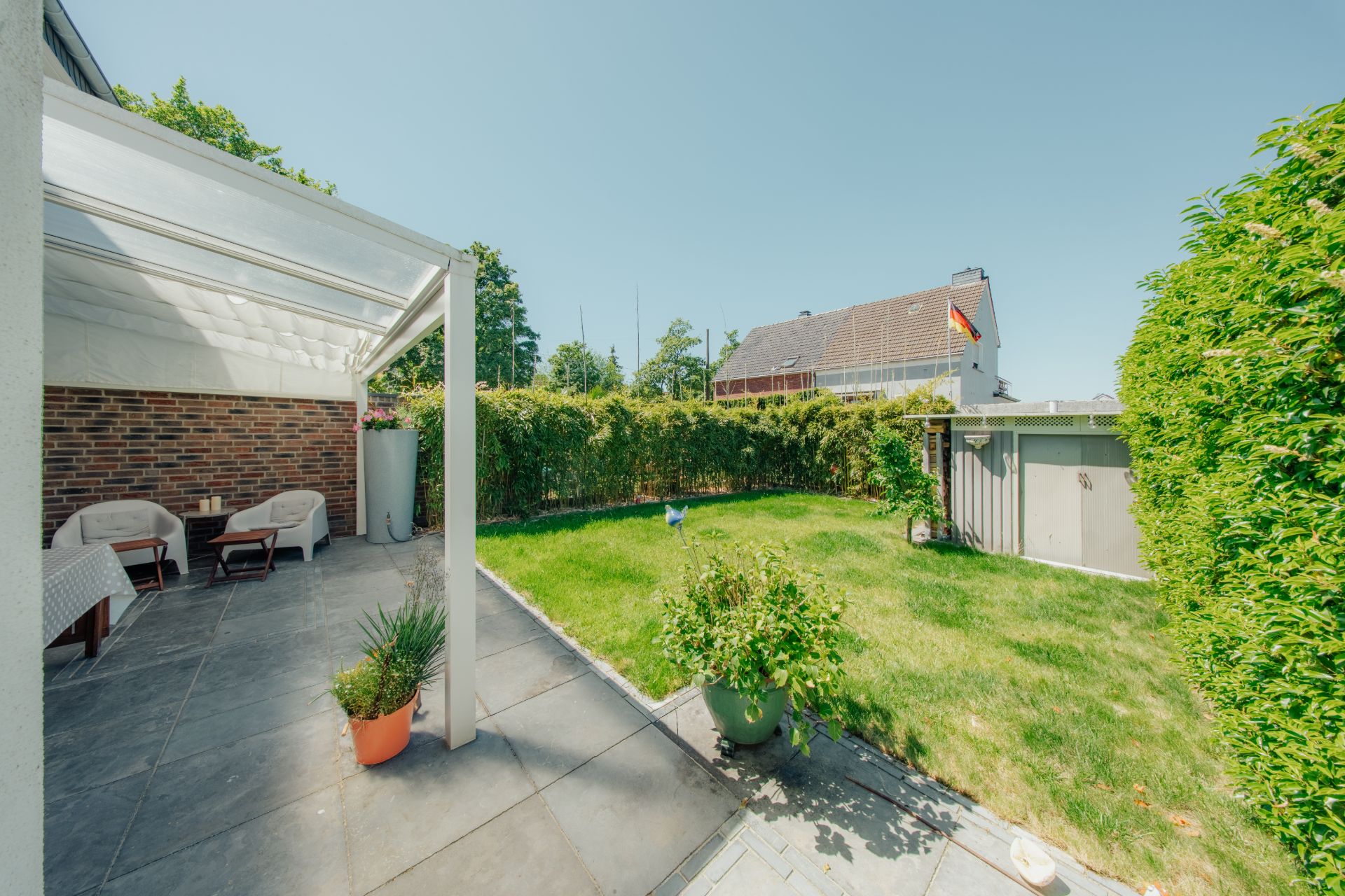 Überdachte Terrasse mit Sitzmöbeln und Blick auf einen grünen Garten mit Hecke und Geräteschuppen.