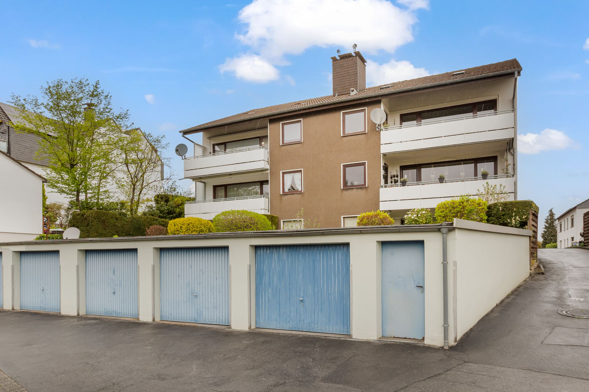 Mehrfamilienhaus mit drei Etagen, Balkonen und blauen Garagentoren im Vordergrund.