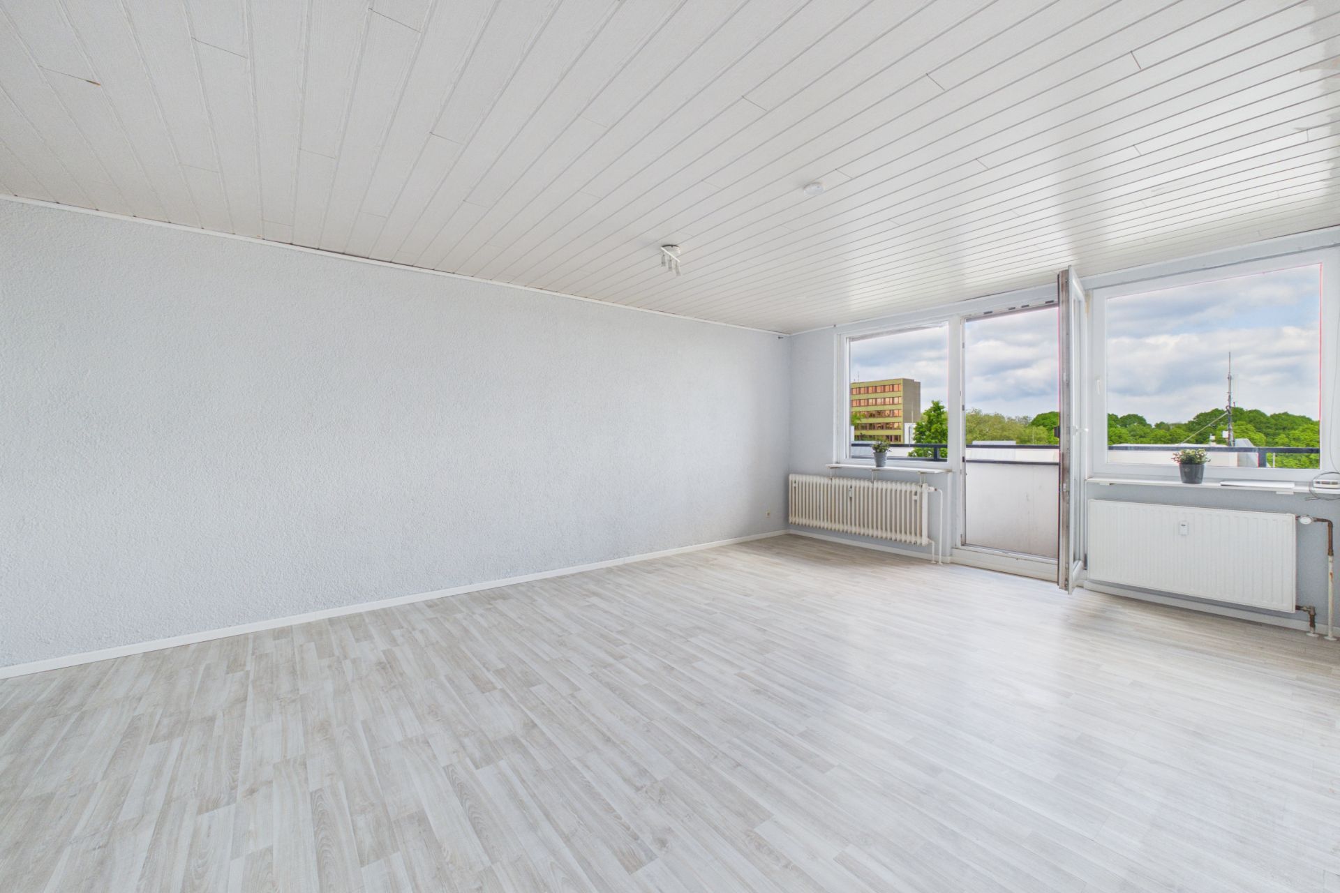 Helles, leeres Zimmer mit weißen Wänden, Parkettboden und großen Fenstern mit Blick ins Grüne.
