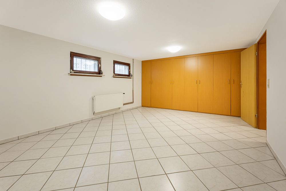Leerer Raum mit weißen Fliesen, zwei kleinen Fenstern und großer Holzschrankwand.