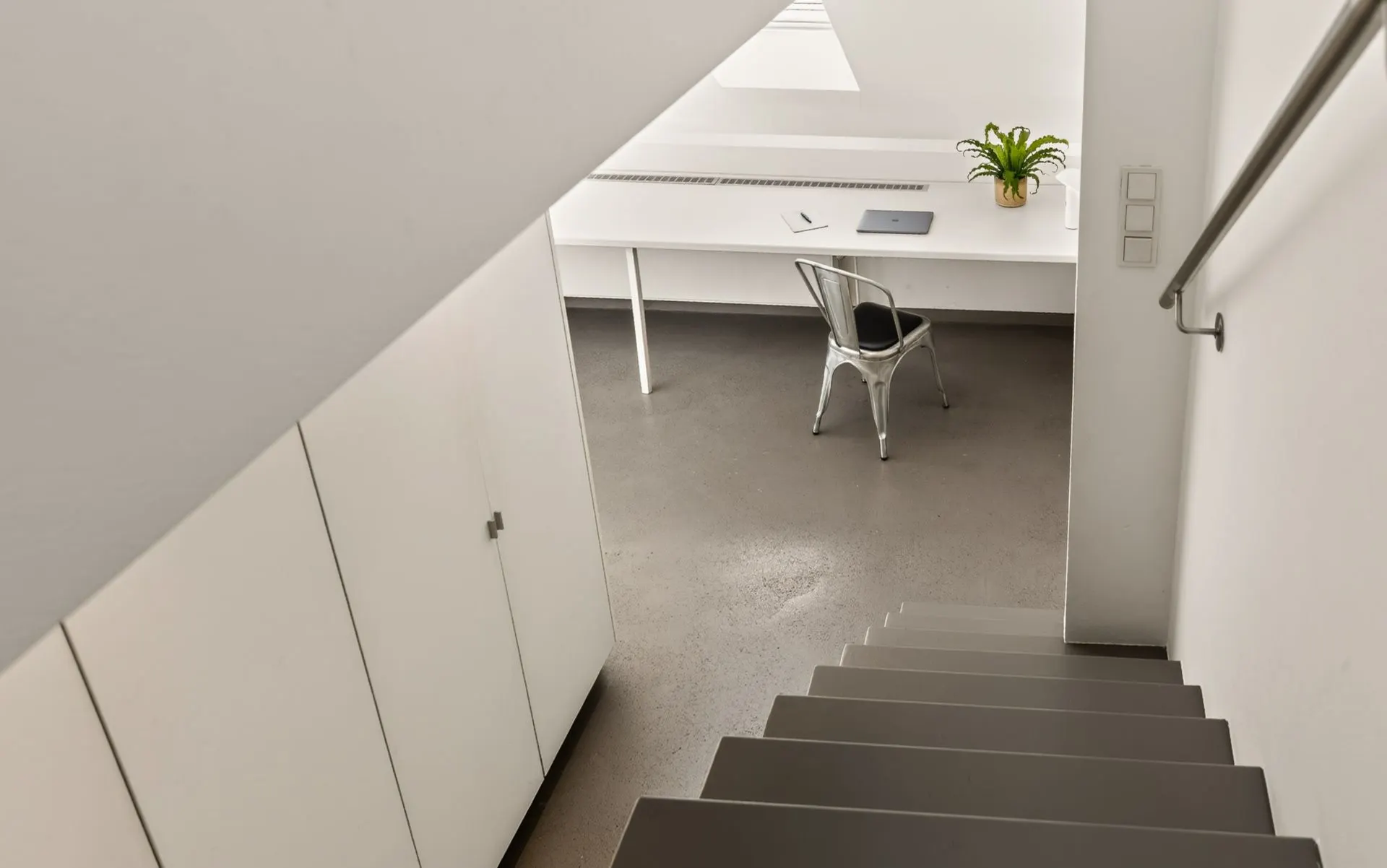 Blick von einer Treppe auf einen Schreibtisch mit Stuhl, Laptop und Pflanze in einem hellen Raum.