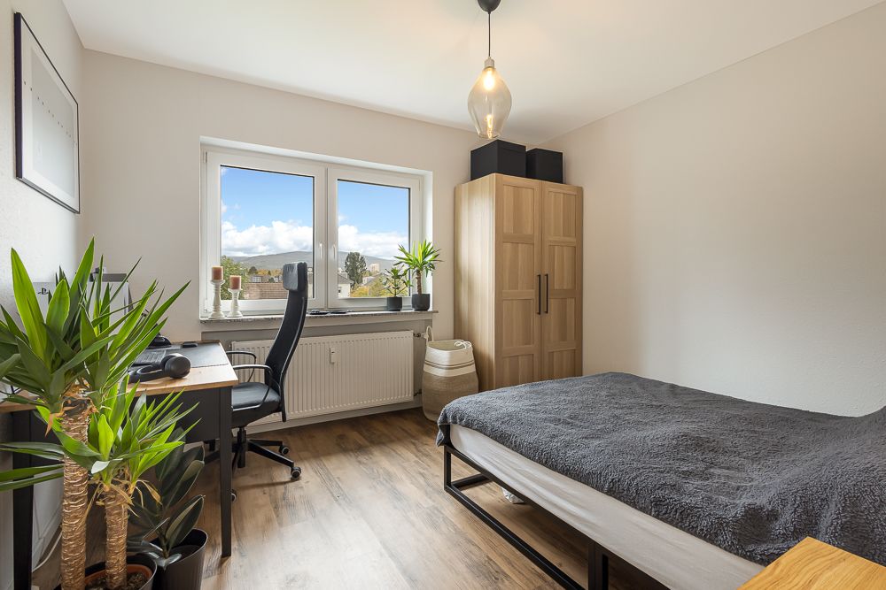Helles Schlafzimmer mit Schreibtisch, Stuhl, Bett, Kleiderschrank und Pflanzen am Fenster.