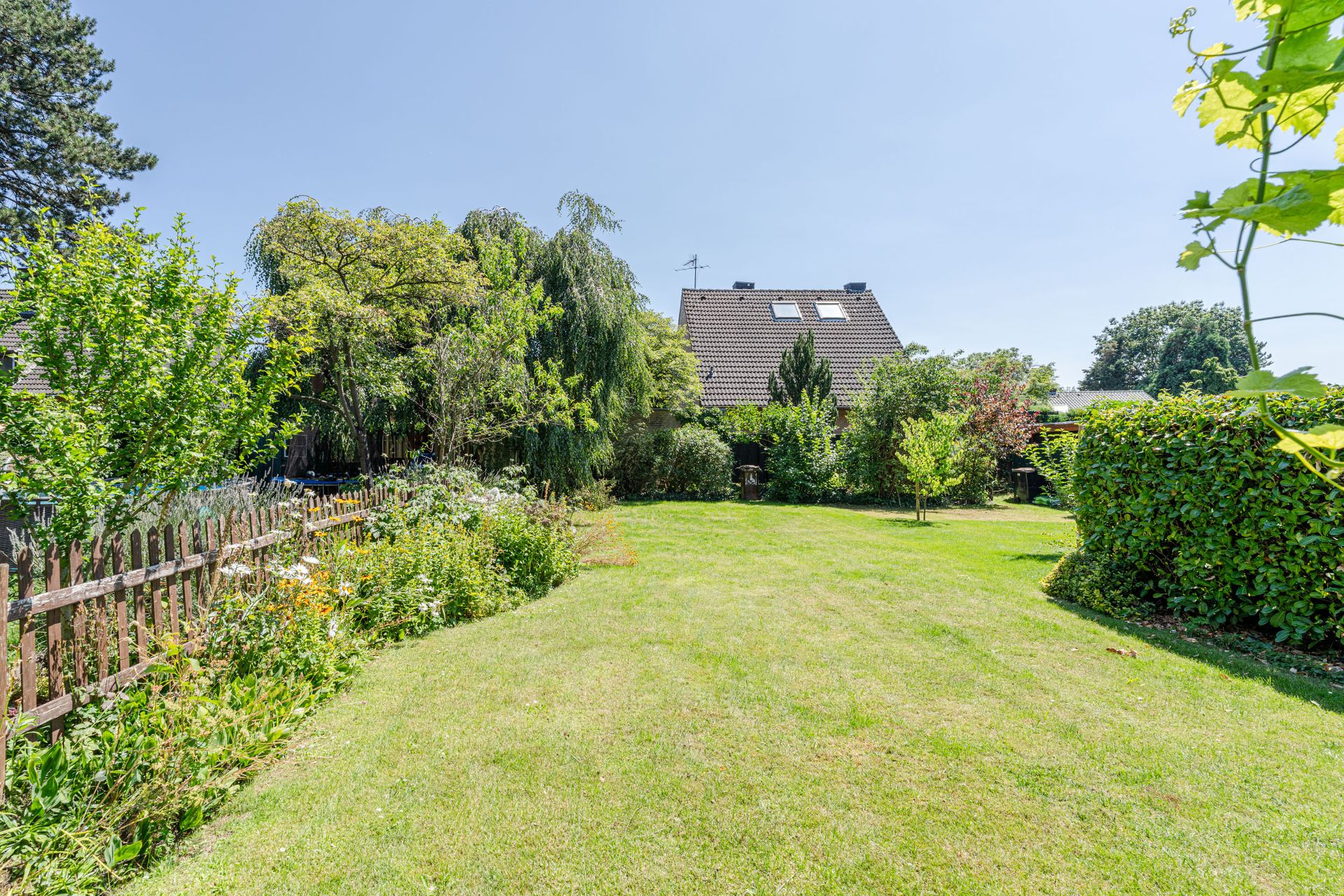 Großer grüner Garten mit Rasen, Bäumen und Blumen, hinten ein Haus mit Schrägdach.