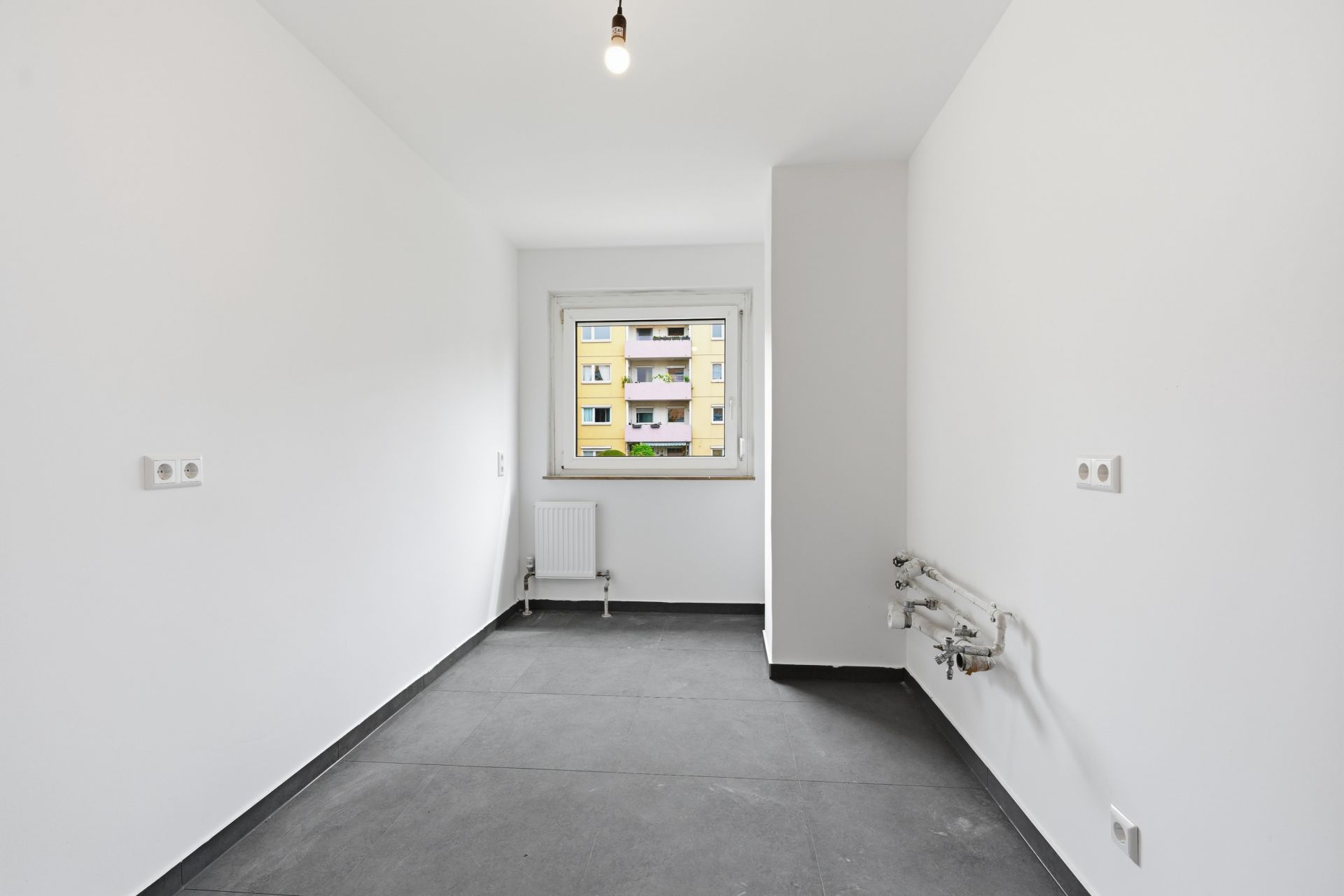 Leerer, weißer Raum mit grauem Boden, Fenster und Heizung, ohne Möbel.