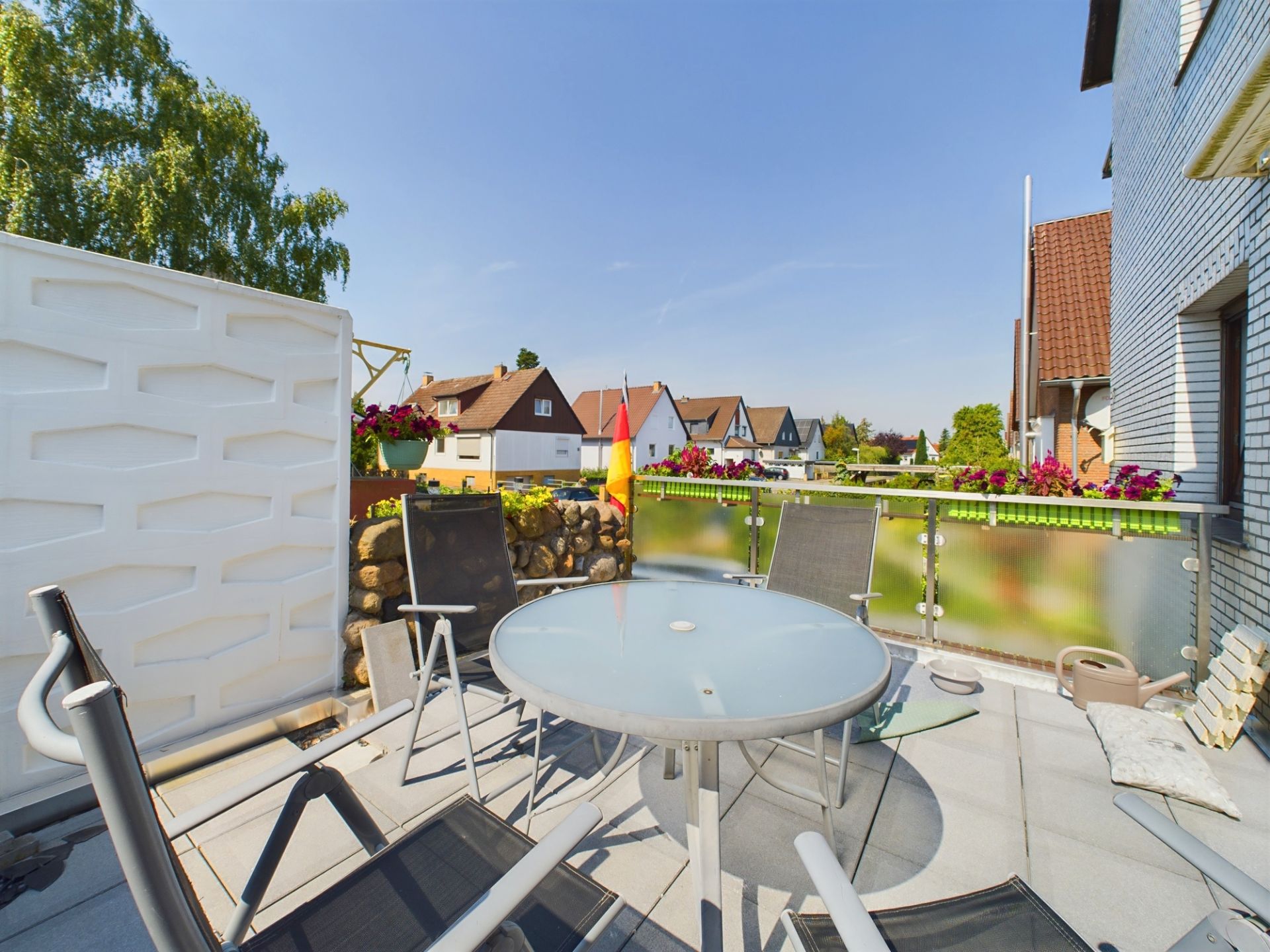 Balkon mit Glastisch, vier Stühlen, Blumenkästen und Blick auf Wohnhäuser.