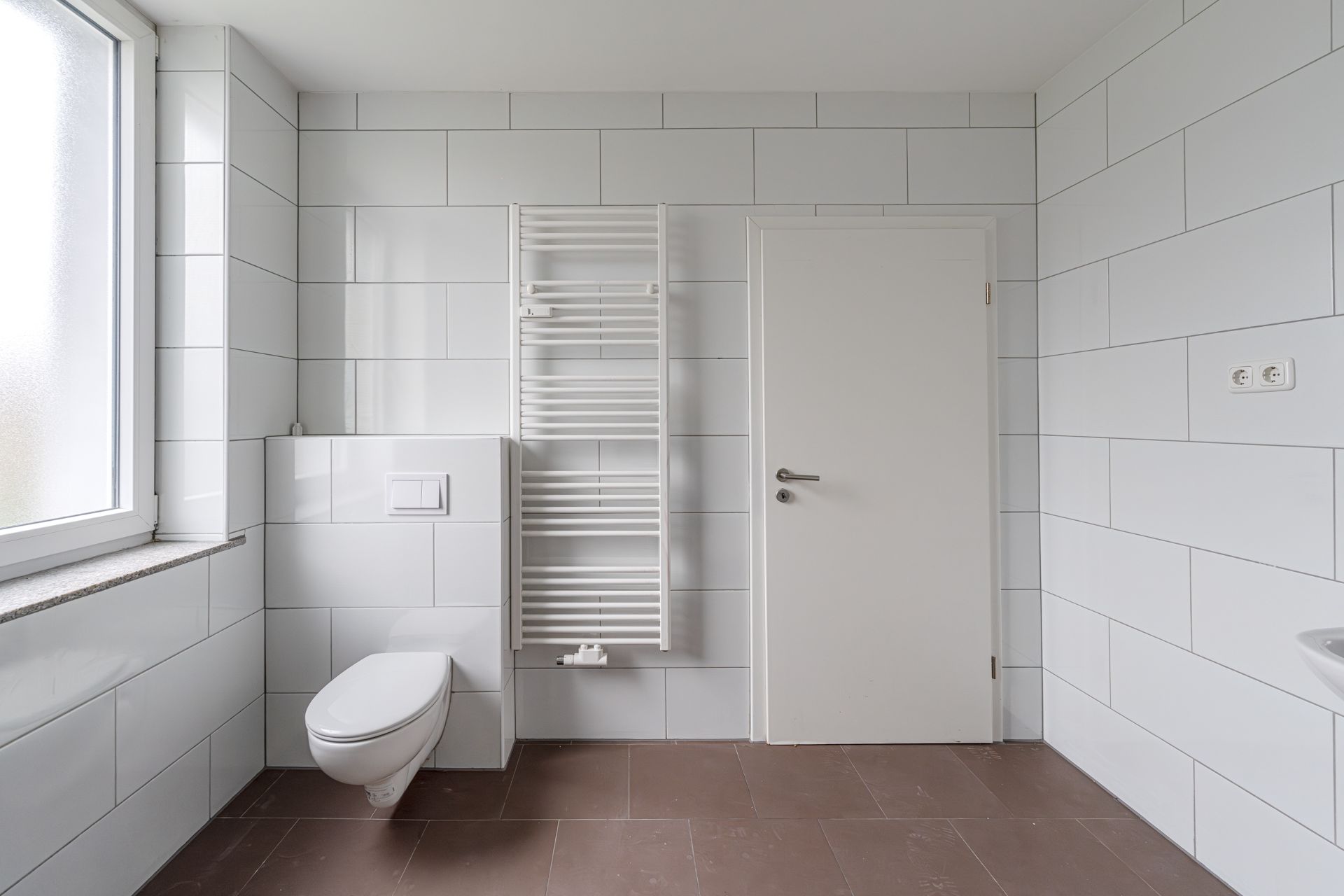 Helles Bad mit wandhängender Toilette, Handtuchheizkörper, Tür und Fenster.