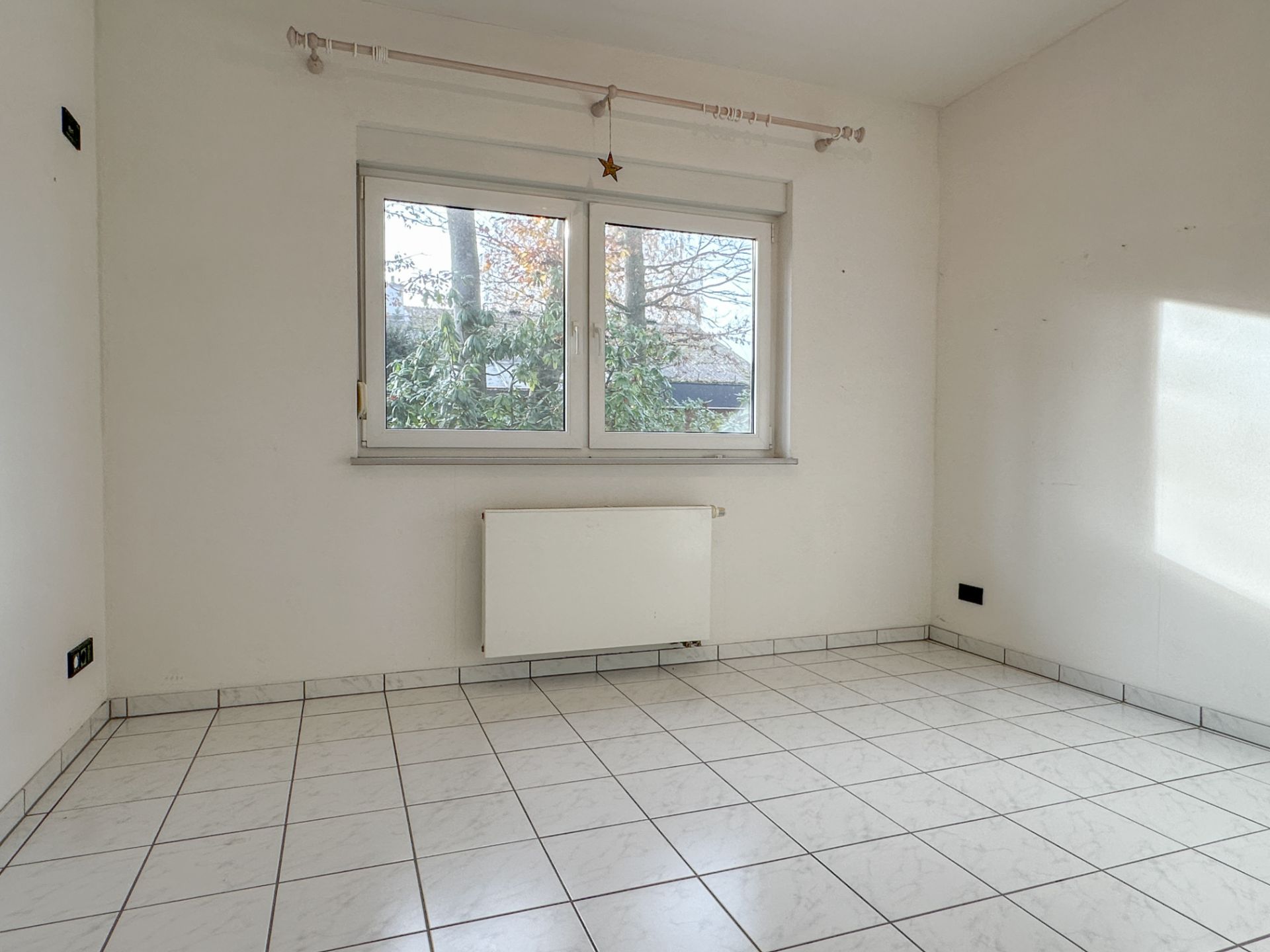 Leeres Zimmer mit weißen Fliesen, Fenster, Heizkörper und Gardinenstange.