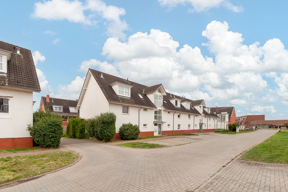 Wohnstraße mit mehreren weißen Reihenhäusern und gepflastertem Weg, grünen Büschen und Wiese.