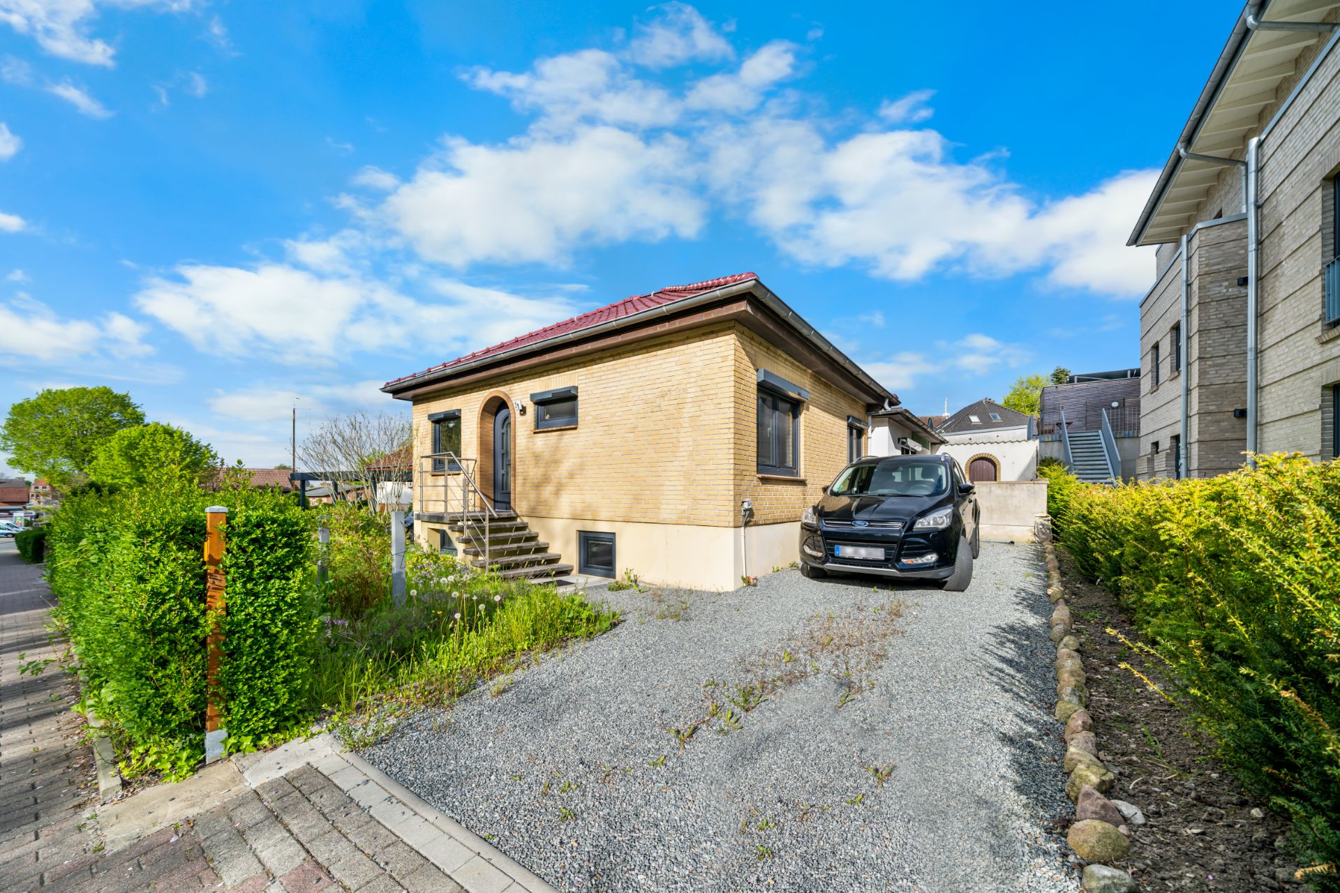 Einfamilienhaus mit rotem Dach, Kiesauffahrt und geparktem Auto, umgeben von Büschen.