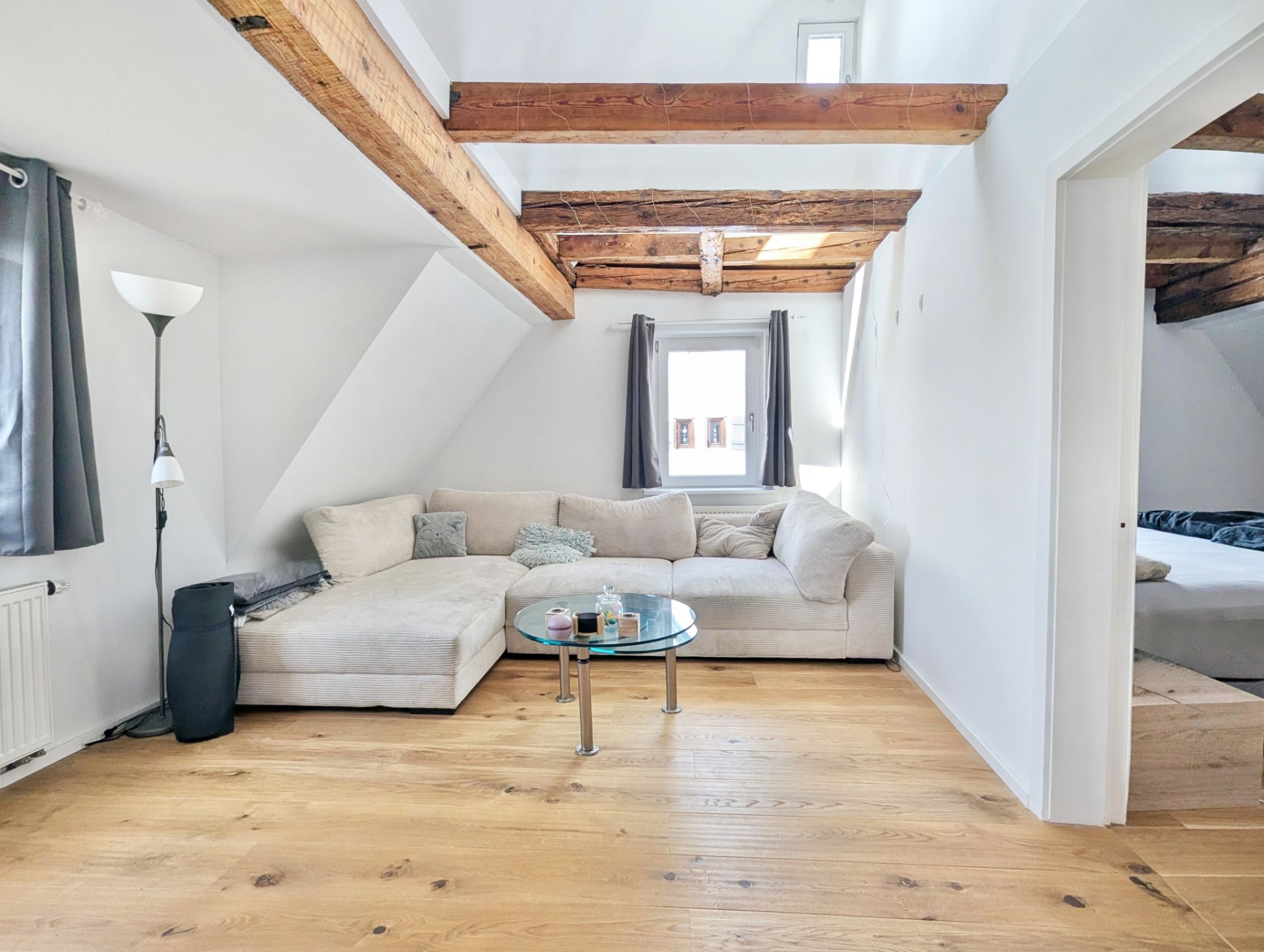 Helles Wohnzimmer mit Holzbalkendecke, Ecksofa, Couchtisch und Fenster mit grauen Vorhängen.