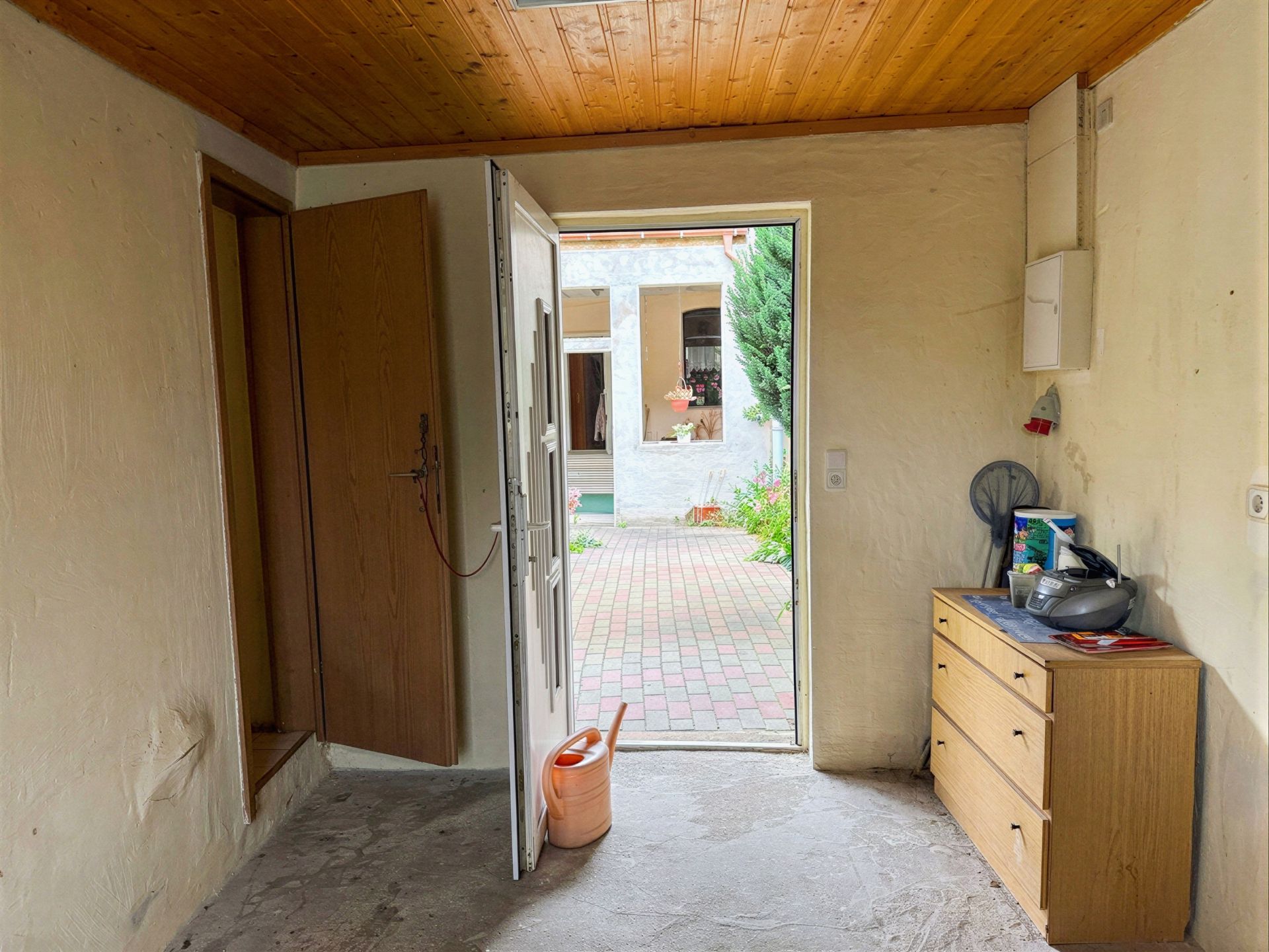 Flur mit Holzboden, Kommode, offener Tür und Blick auf einen Innenhof mit Pflastersteinen.