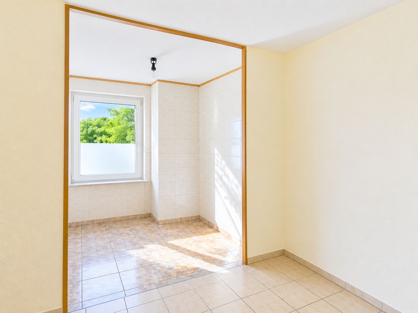 Heller, leerer Raum mit Fliesenboden und großem Fenster, durch Türöffnung sichtbar.