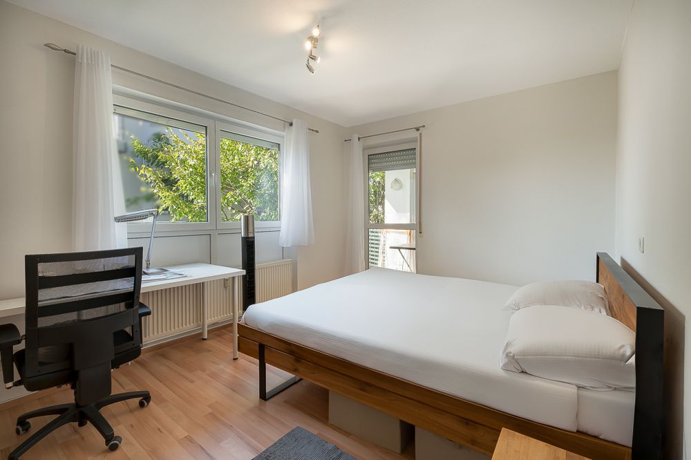 Helles Schlafzimmer mit Doppelbett, Schreibtisch, Bürostuhl und großen Fenstern mit Blick ins Grüne.