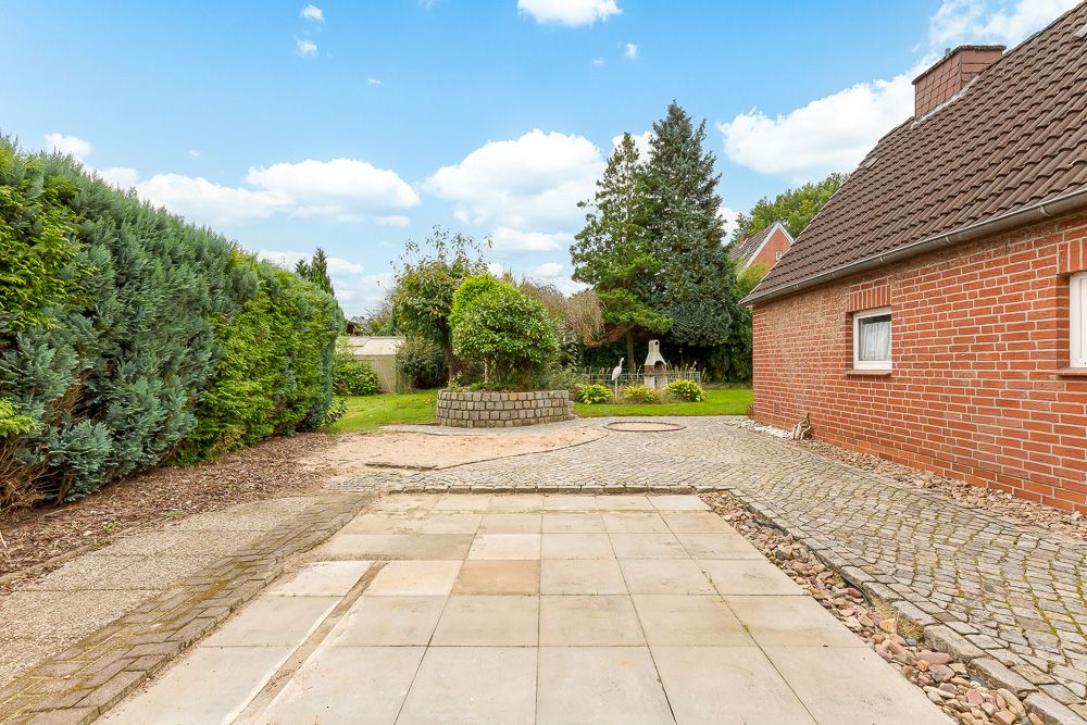 Gepflasterter Hof neben einem roten Backsteinhaus mit Garten und grünen Büschen.