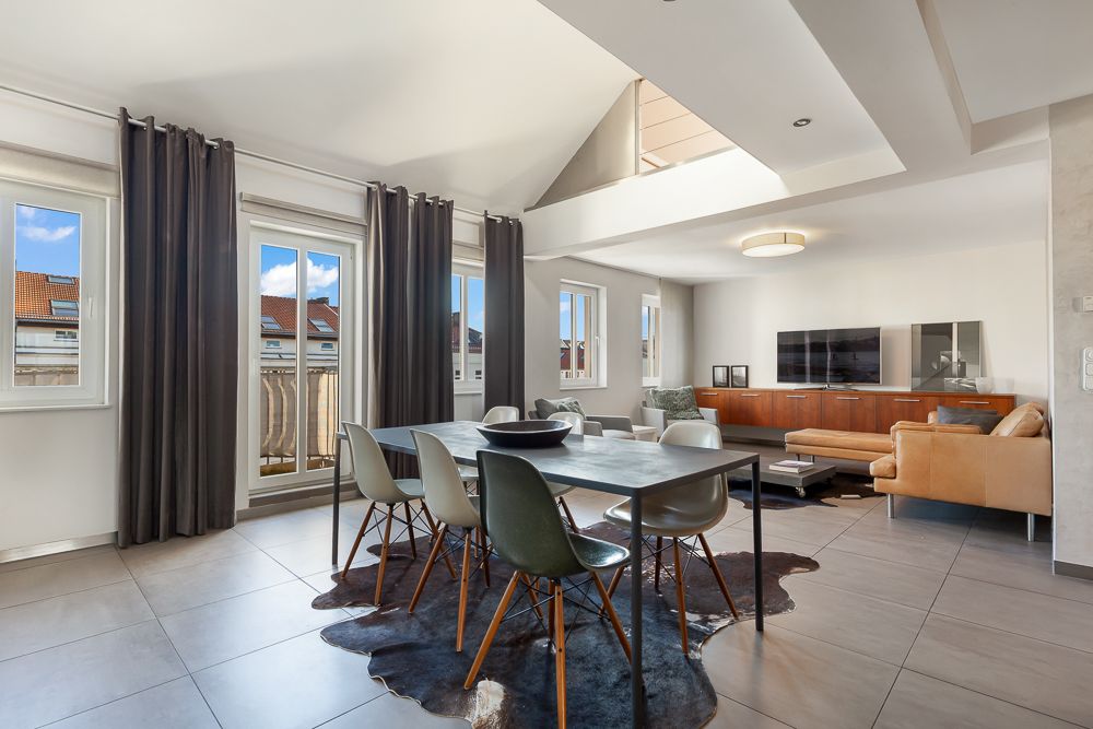Helles Wohnzimmer mit Esstisch, Stühlen, Sofa und TV, große Fenster, Fliesenboden.