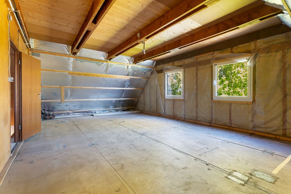 Leerer Dachboden mit offenliegenden Holzbalken, zwei Fenstern und sichtbarer Isolierung.