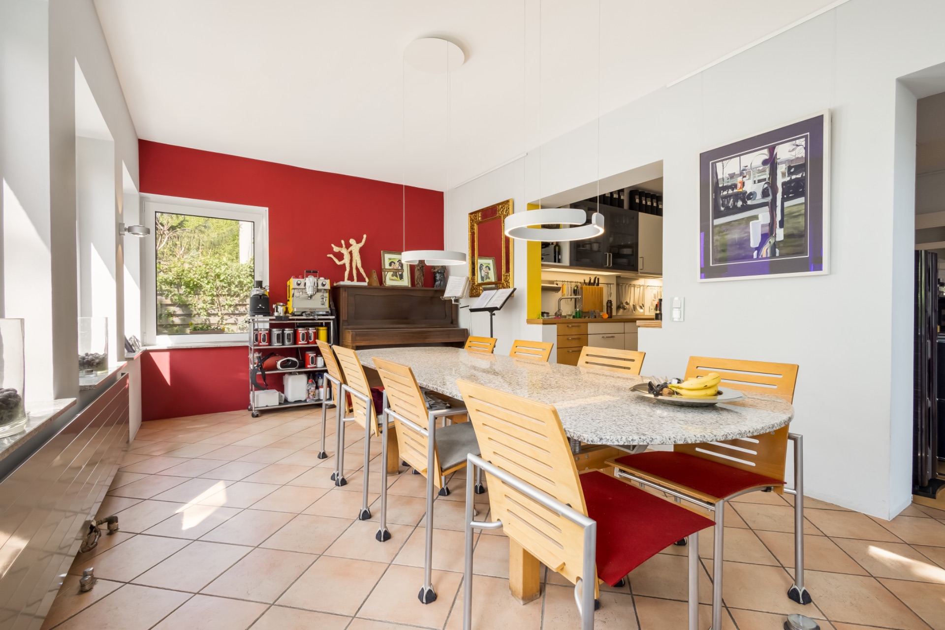 Esszimmer mit großem Tisch, Holzstühlen, roter Wand, Fenster und offenem Zugang zur Küche.