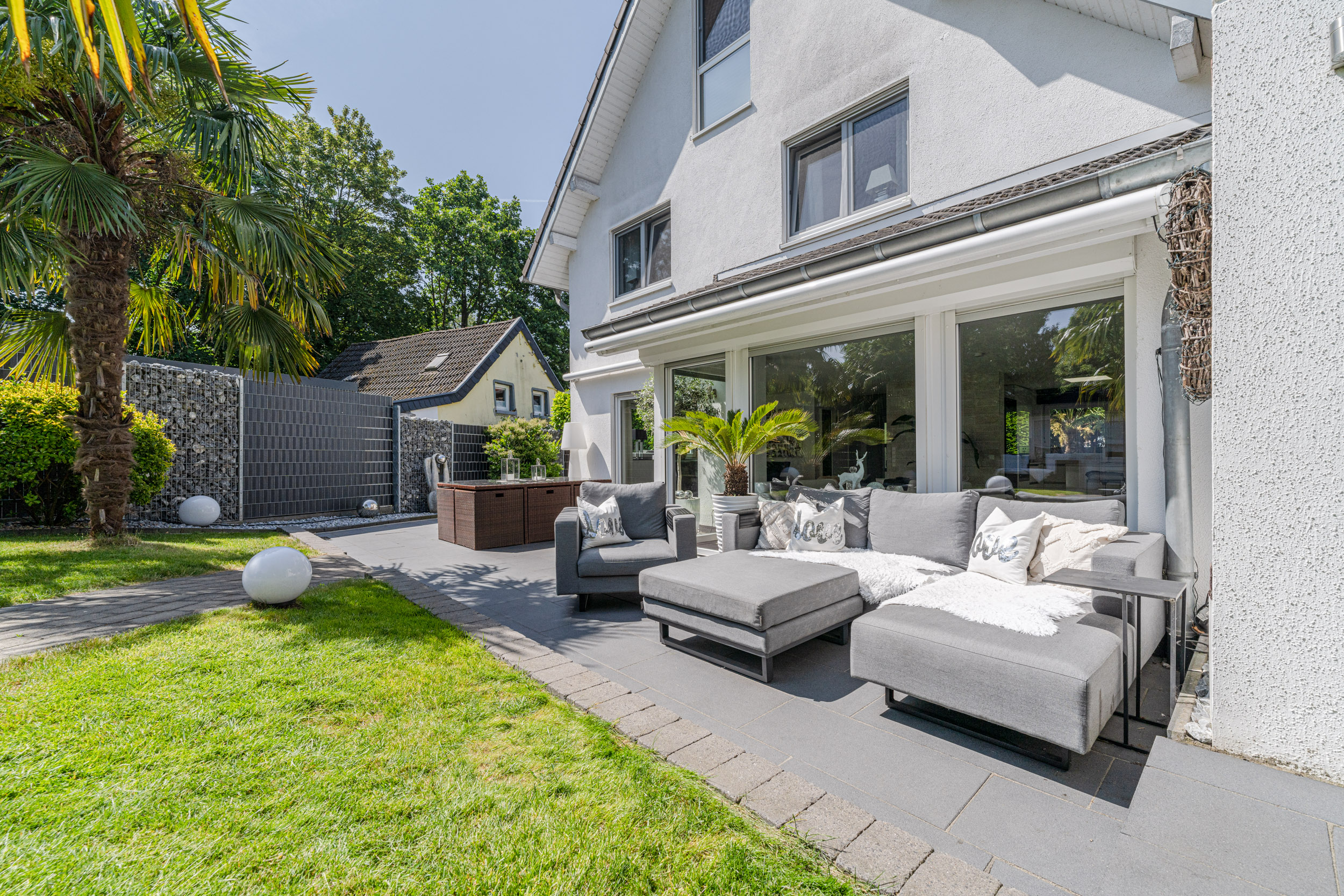 Moderne Terrasse mit grauem Sofa, Palmbaum und Blick auf weißen Wohnhausgarten.